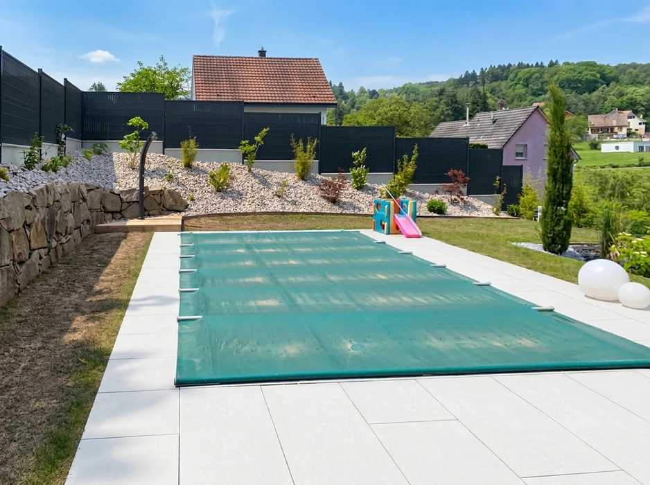 Jardin résidentiel extérieur avec piscine rectangulaire couverte d'une bâche hivernage verte à sangles, posée sur margelles en carrelage grès cérame blanc